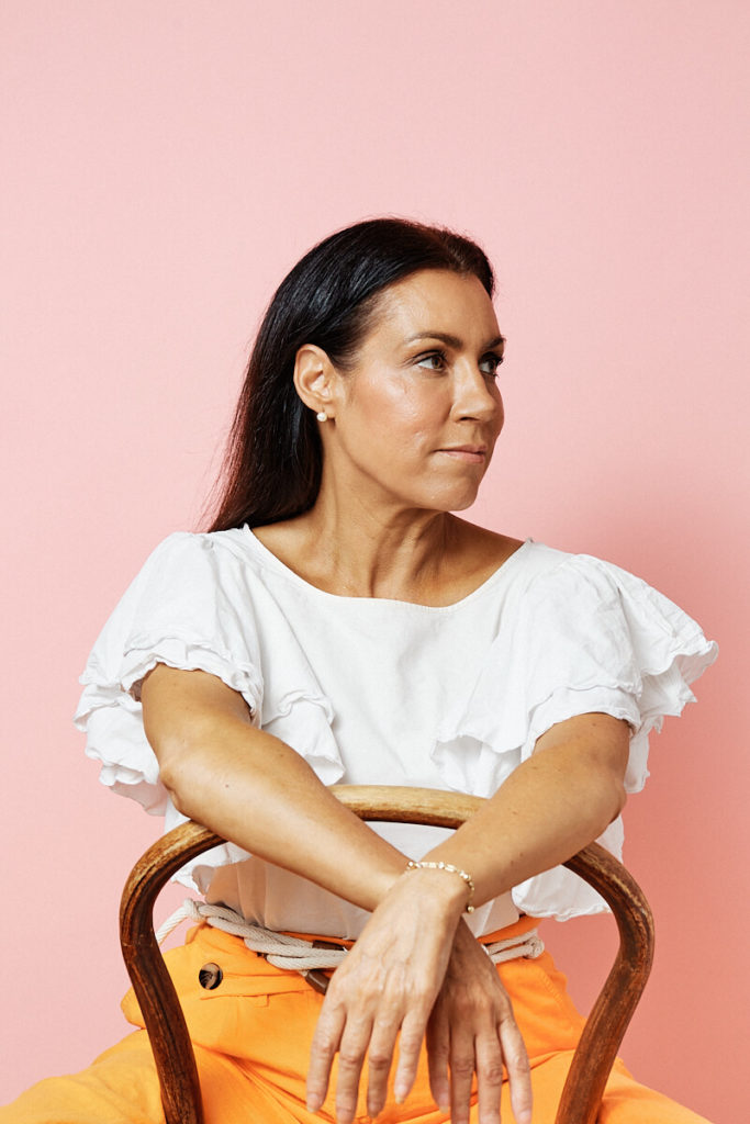 Studio headshot by Photoform*.  Margie is wearing orange pants, a white short sleeved shirt with frills.  She is straddling a wooden chair.  The background is pink and Margie has a determined expression.