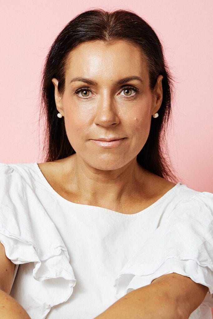 Studio headshot of Margie Thomas by Photoform*.  Margie is wearing a white short sleeved shirt with frills.  Her hair is dark and long and brushed back. Pink background and Margie has a determined expression.