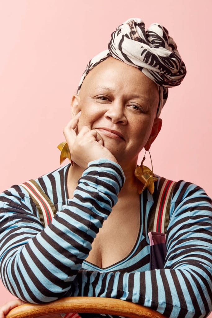 Studio headshot by Photoform*. Woman with a patterned turban and a striped blue and black top against a pink background.  The woman has her hand on her chin.