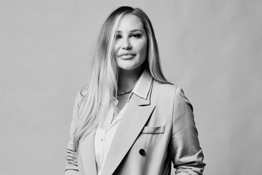 Black and white studio headshot by Photoform*. She is wearing a 3/4 length coat with a white shirt.