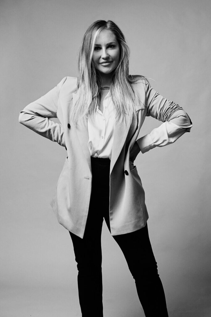 Black and white studio headshot by Photoform* of woman with hands on hips. She's wearing a 3/4 length coat with a white shirt and black trousers.