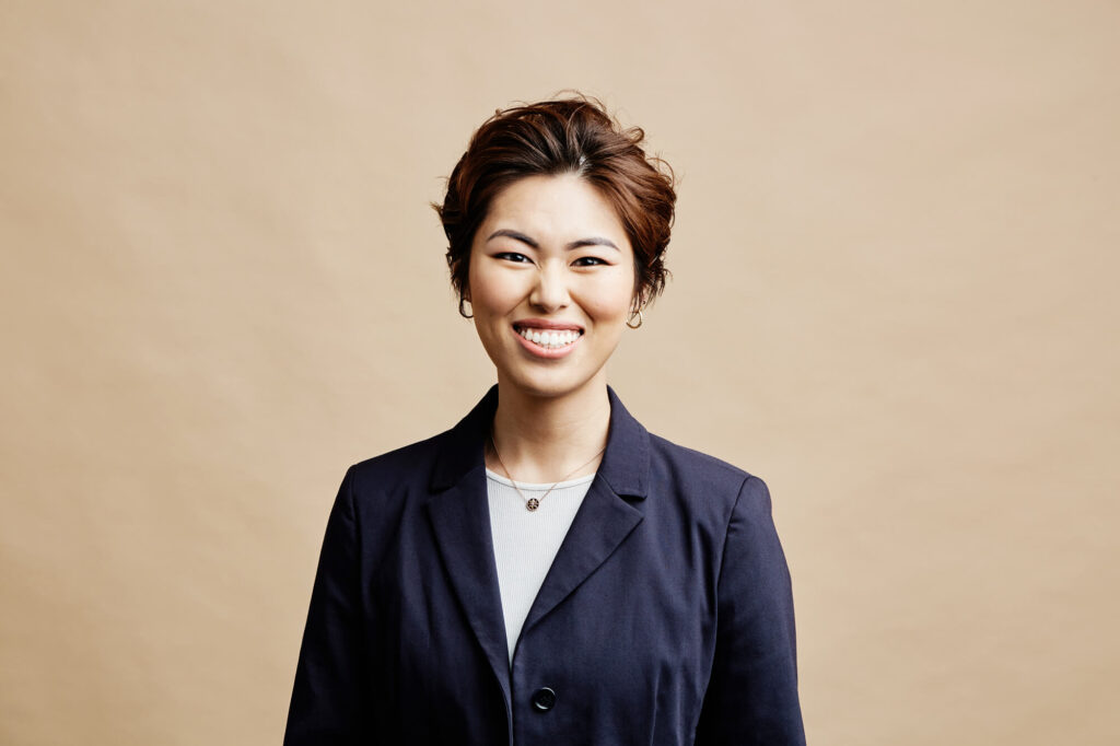 Studio headshot by Photoform* of woman smiling wearing a blue jacket against cream background.