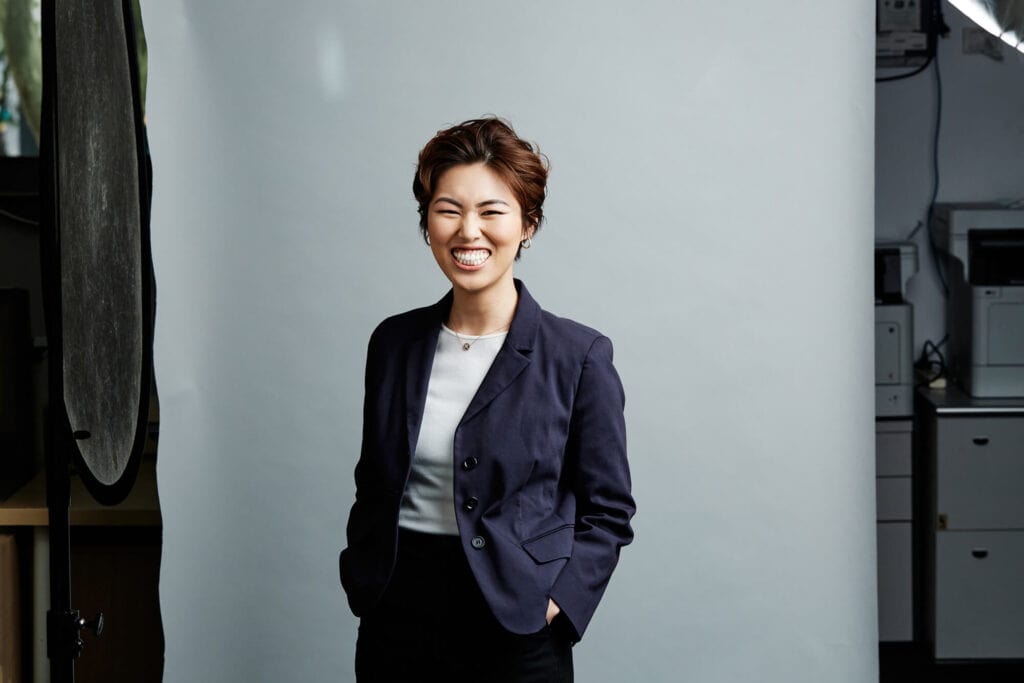 Studio headshot by Photoform* of woman smiling wearing a blue jacket against light grey background with office behind the background.