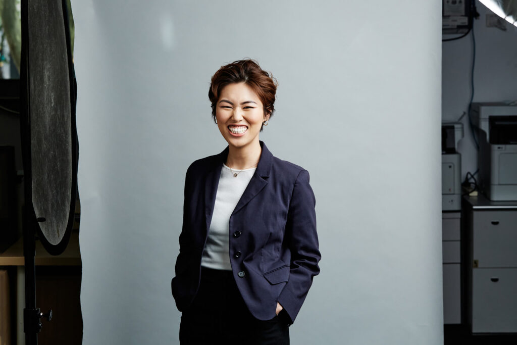 Studio headshot by Photoform* of woman smiling wearing a blue jacket against light grey background with office behind the background.