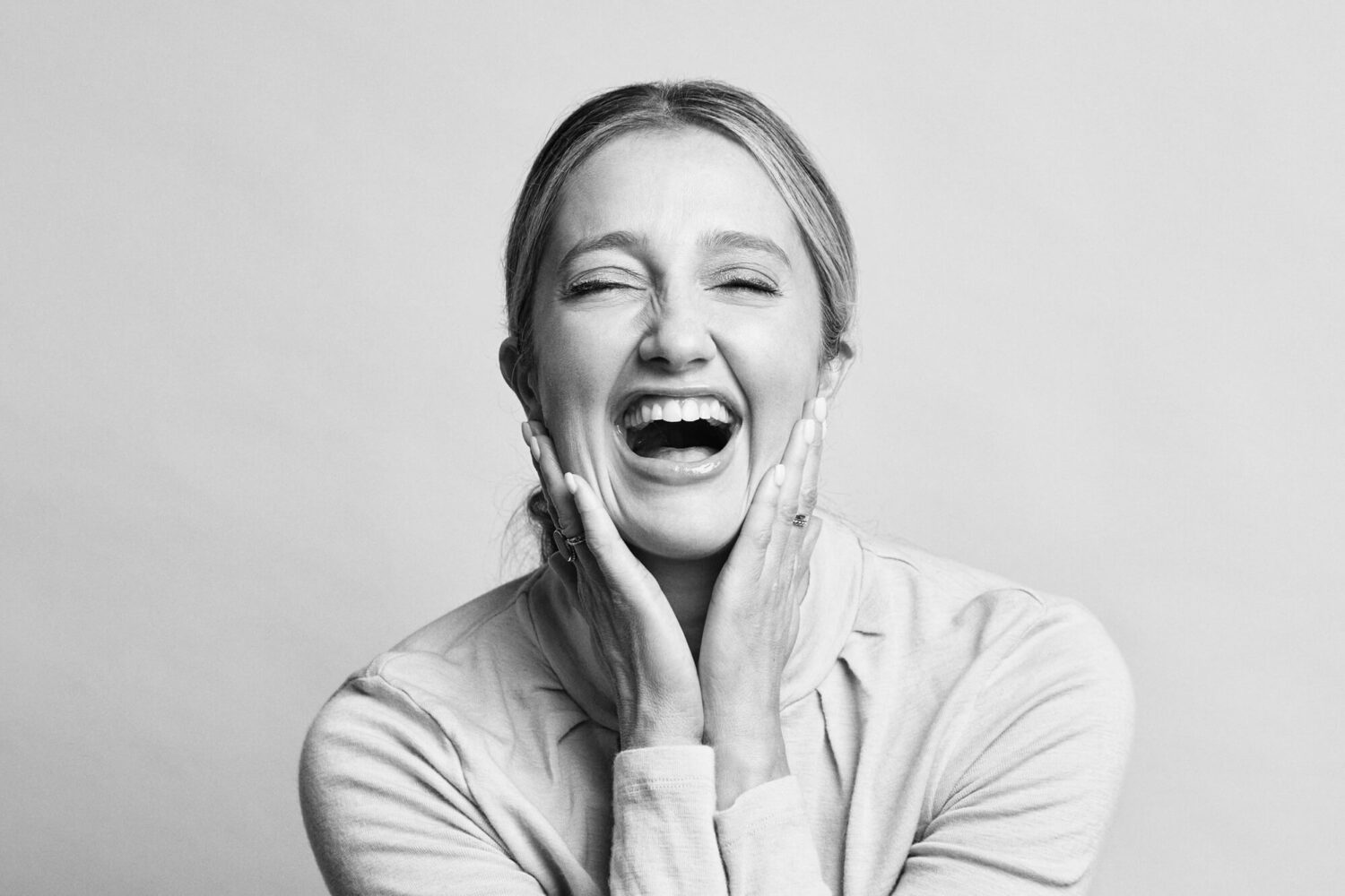 Black and white portrait.  A blonde woman is laughing with both palms against either side of her face.