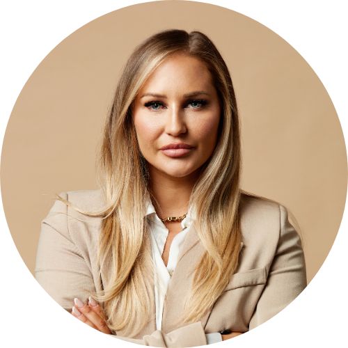 A LinkedIn profile picture with a fashion edge.
She is wearing a camel coloured coat, white shirt and has a determined expression.
She's against a beige coloured background.
