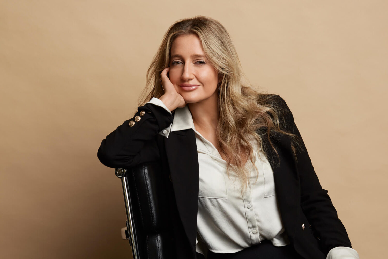 Blonde woman on a black leather chair with her chin resting on the palm of her hand.
She is wearing a white shirt and black jacket.  She is in a studio against a beige coloured background.
