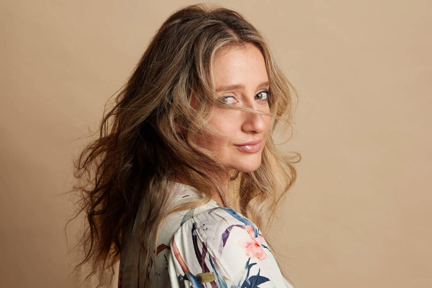 Blonde woman headshot in a studio against a beige background.
Her head is turned towards the camera and her hair is swept across her face.  She's peering through her hair.  She's wearing a floral dress.