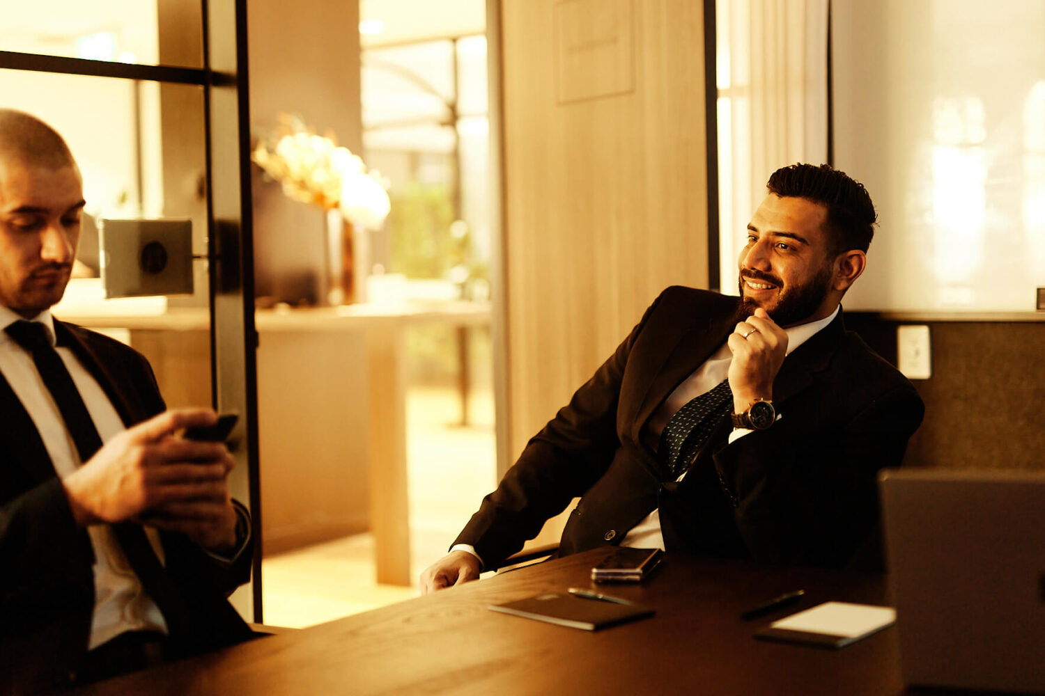 2 men are in a meeting.  Both are in suits and one is looking at his phone.
It's golden hour at sunset and the meeting room is filled with golden light. Photo by Photoform*.