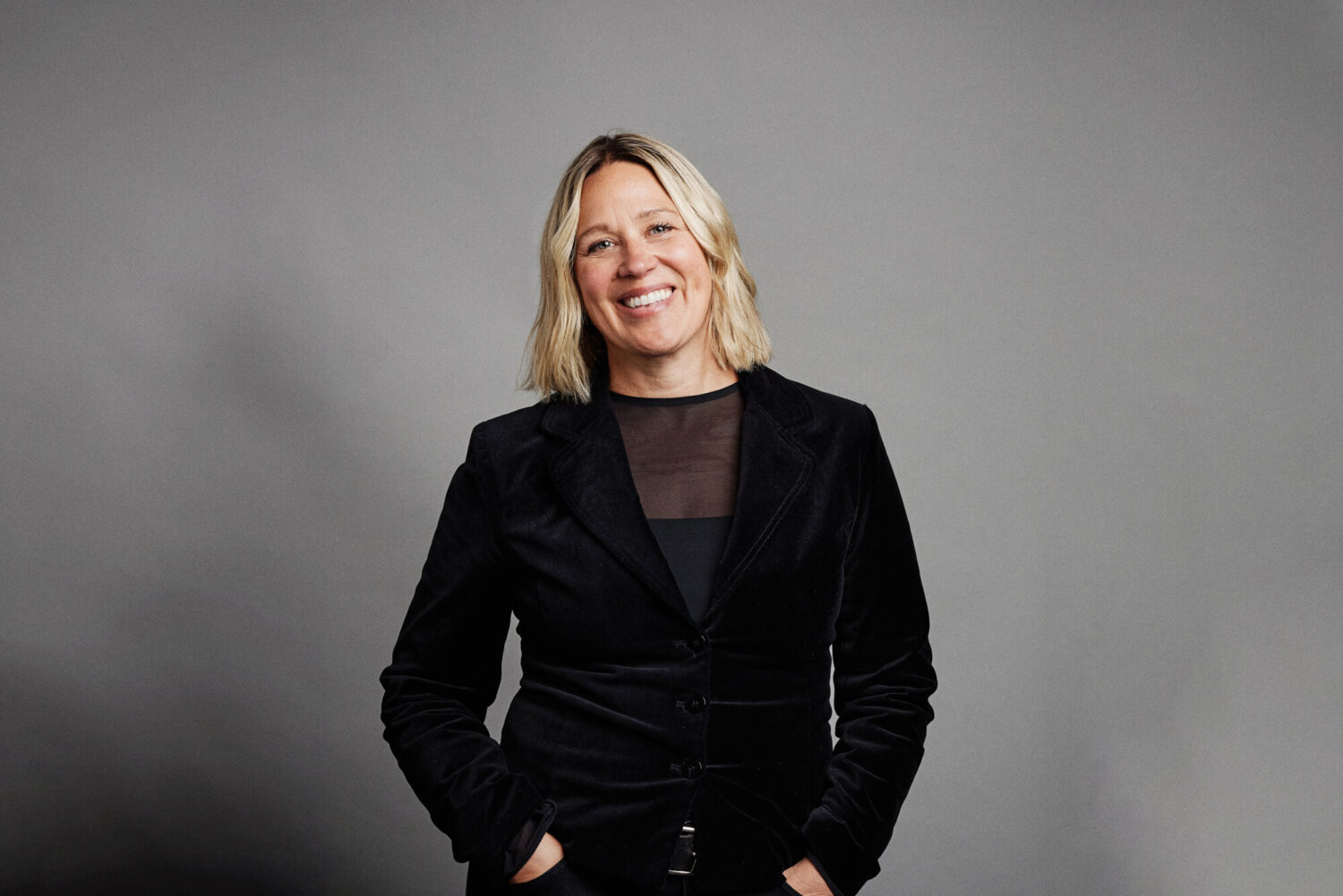 Corporate headshot by Photoform*. Smiling blonde woman wearing all black against a grey background.