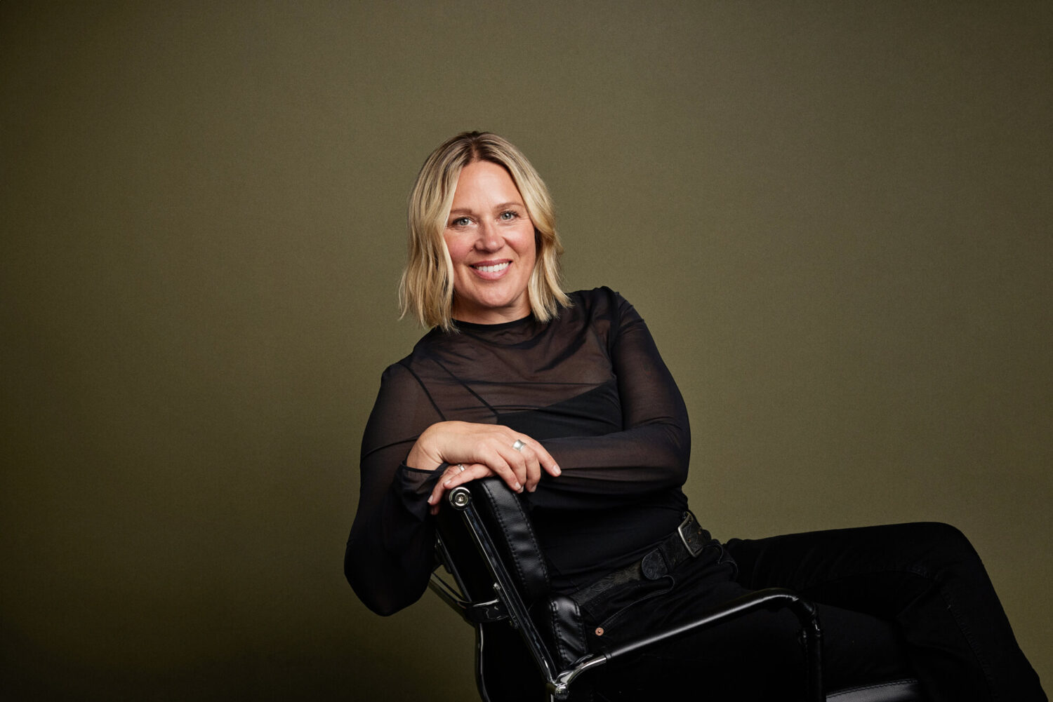 Portrait by Photoform*. Woman is smiling at the camera and she's sitting on a leather black chair.
She's wearing all black and has blonde hair.  She's against an olive coloured background/
