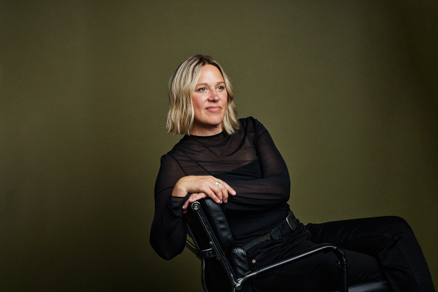 Portrait by Photoform*. Woman is looking to the right and she's sitting on a leather black chair.
She's wearing all black and has blonde hair.  She's against an olive coloured background/