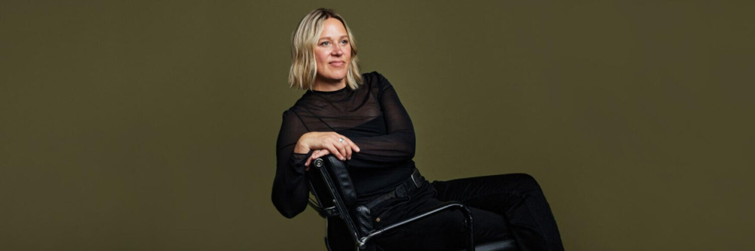 Example of an olive background used in studio photoshoots. Woman is sitting on a leather and chrome chair and wearing all black. All headshots are by Photoform*.