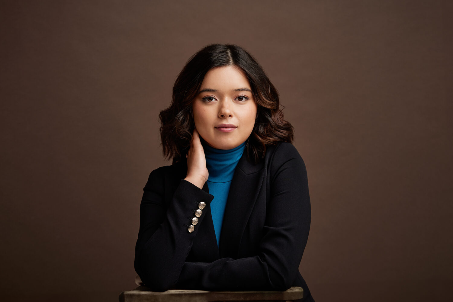 Corporate headshot of Steph by Photoform*.
Steph is wearing a blue turtleneck and black jacket and is against a dark brown background.