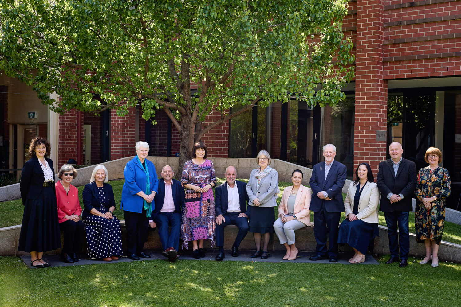 Group photo of the Mercy Ministry Companions at the 2024 Annual Conference, seated and standing outdoors in front of a tree and red brick building. This image illustrates tips for composing large group photos.