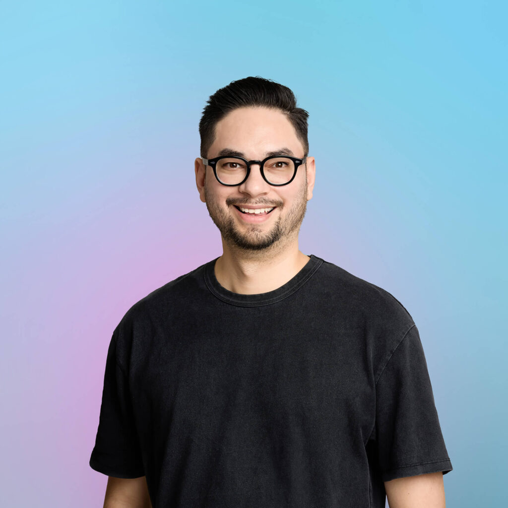 Nicholas Carlton, Spoony CEO. Nicholas has glasses, is wearing a black tshirt and is smiling against a pink and blue gradient.